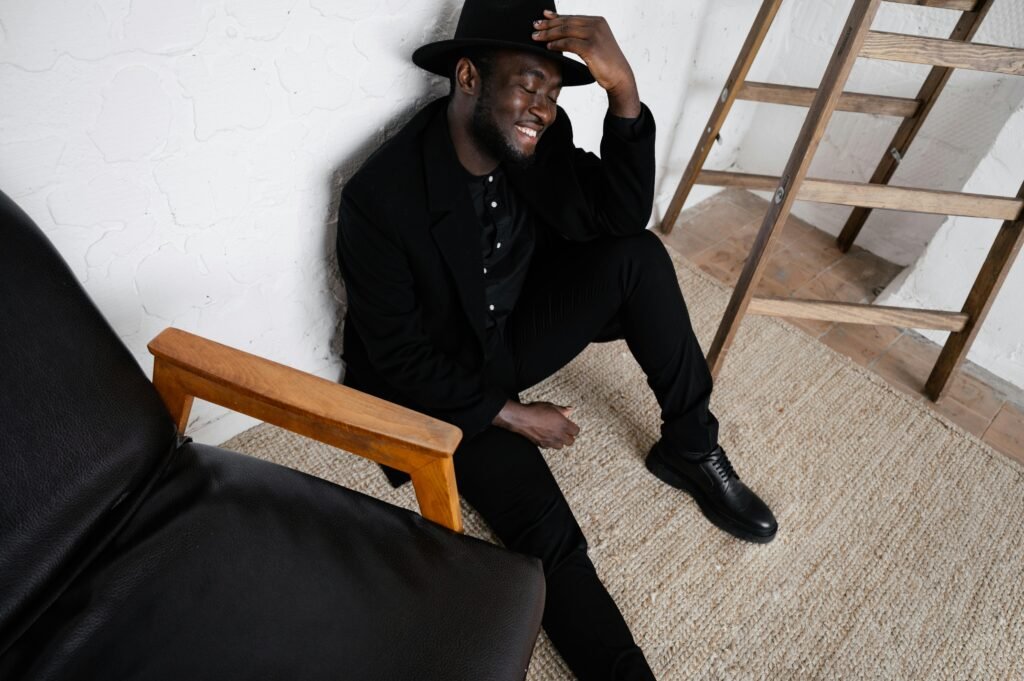 A stylish man in black attire smiling while sitting on the floor indoors, wearing a hat.