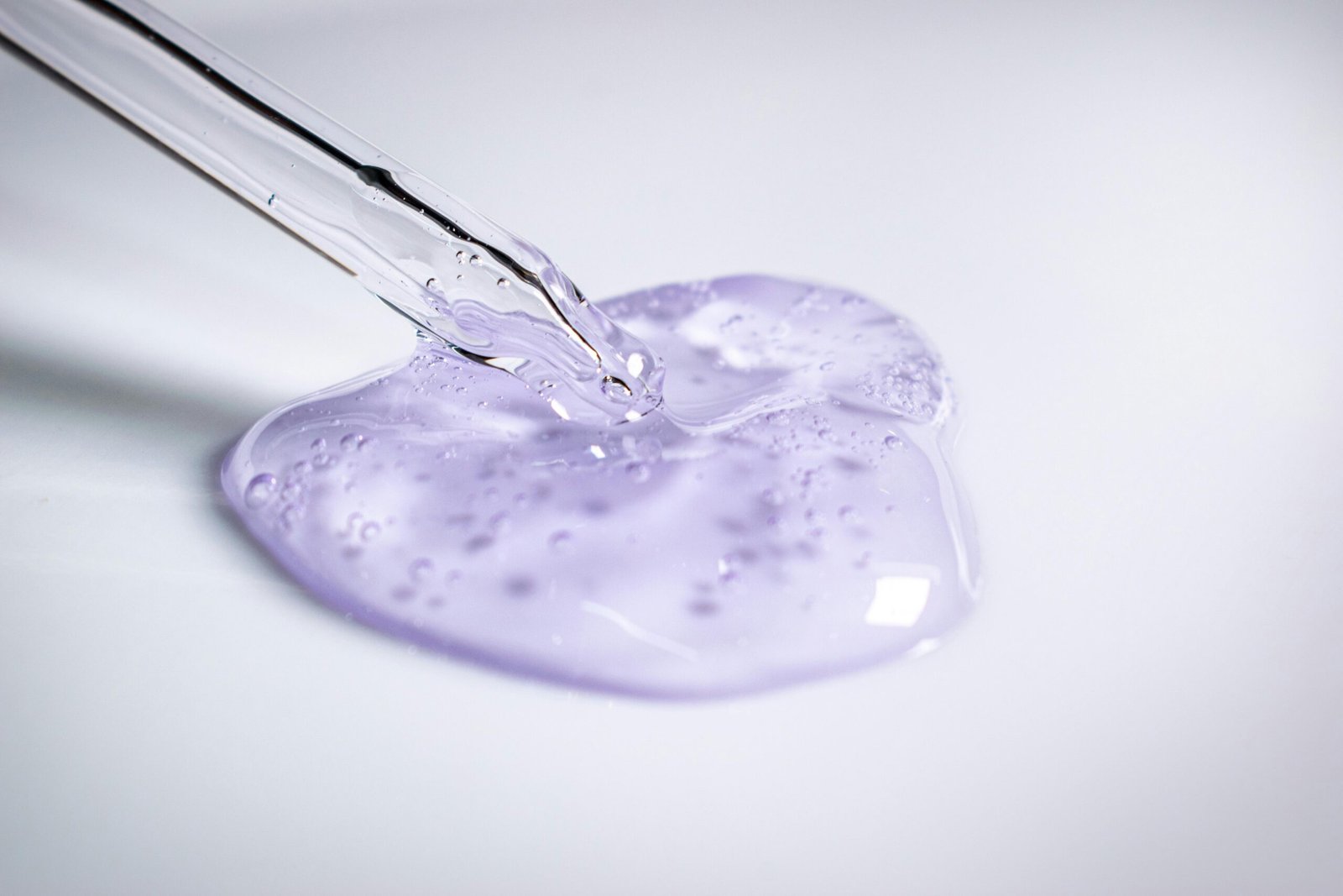 A pipette dispensing clear purple cosmetic gel onto a smooth surface.