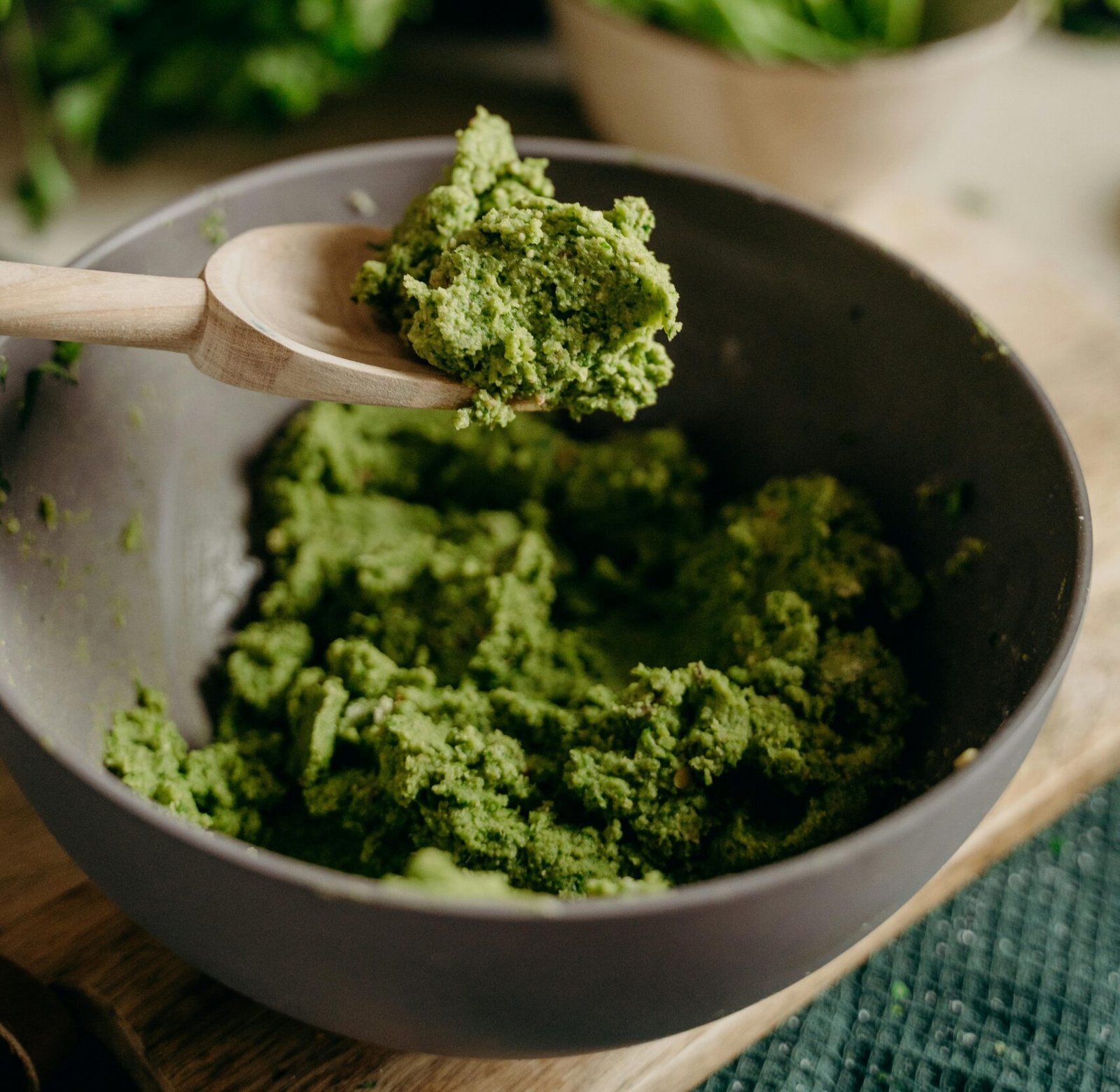 A close-up of fresh green pesto in a ceramic bowl with a wooden spoon, set in a rustic kitchen.